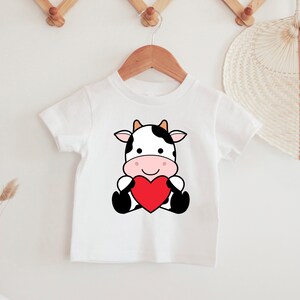 K&ouml;nnte beinhalten: Wei&szlig;es Kinder-T-Shirt mit einer Cartoon-Kuh, die ein rotes Herz h&auml;lt. Die Kuh hat schwarz-wei&szlig;e Flecken, eine rosa Nase und einen l&auml;chelnden Gesichtsausdruck. Das T-Shirt h&auml;ngt an einem Holzb&uuml;gel.