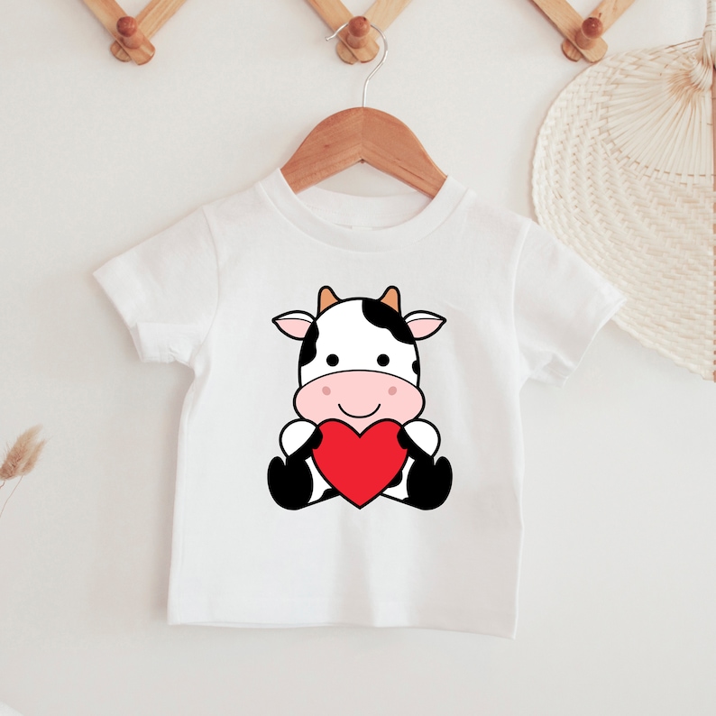 K&ouml;nnte beinhalten: Wei&szlig;es Kinder-T-Shirt mit einer Cartoon-Kuh, die ein rotes Herz h&auml;lt. Die Kuh hat schwarz-wei&szlig;e Flecken, eine rosa Nase und einen l&auml;chelnden Gesichtsausdruck. Das T-Shirt h&auml;ngt an einem Holzb&uuml;gel.