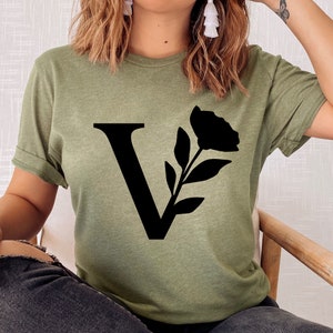 Pu&ograve; includere: T-shirt verde oliva con una grande "V" nera e un design floreale. La grafica include un fiore e foglie. La maglietta ha maniche corte e una vestibilit&agrave; rilassata.