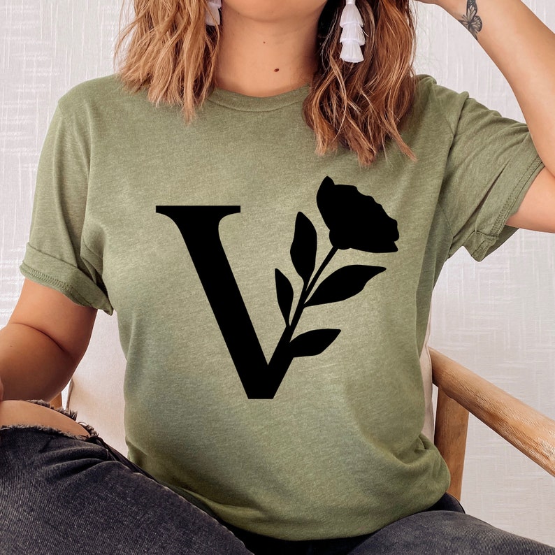 Pu&ograve; includere: T-shirt verde oliva con una grande "V" nera e un design floreale. La grafica include un fiore e foglie. La maglietta ha maniche corte e una vestibilit&agrave; rilassata.