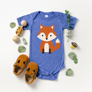 May include: A blue baby bodysuit with a cartoon fox illustration. The fox is orange, white, and brown. The image also includes brown leather baby shoes, a wooden rattle, and a pacifier, suggesting a baby clothing and accessories theme.