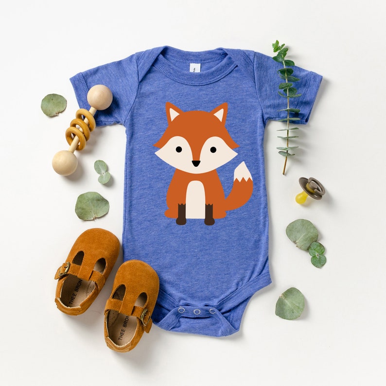 May include: A blue baby bodysuit with a cartoon fox illustration. The fox is orange, white, and brown. The image also includes brown leather baby shoes, a wooden rattle, and a pacifier, suggesting a baby clothing and accessories theme.