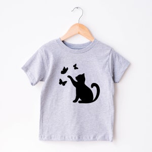 May include: A light gray t-shirt featuring a black silhouette of a cat reaching for three black butterflies. The shirt is hanging on a wooden hanger against a white background. The design is simple and playful.