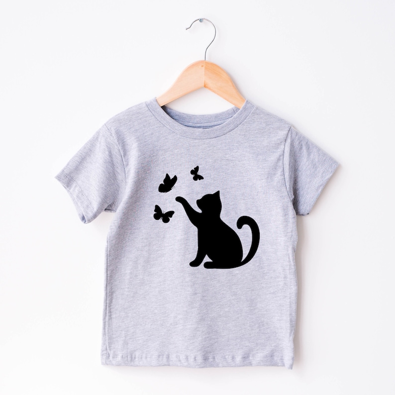 May include: A light gray t-shirt featuring a black silhouette of a cat reaching for three black butterflies. The shirt is hanging on a wooden hanger against a white background. The design is simple and playful.