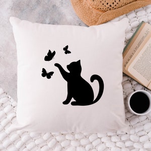 May include: A square, off-white throw pillow featuring a black silhouette of a cat reaching for three black butterflies. The pillow is on a white, textured surface, with a straw hat, open book, and a cup of coffee in the background.