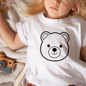Może przedstawiać: Biały t-shirt z kr&oacute;tkim rękawem z czarnym obrysem głowy misia. Miś ma okrągłe czarne oczy i mały czarny nos. Koszulka jest noszona przez dziecko. Widoczny jest drewniany aparat fotograficzny.