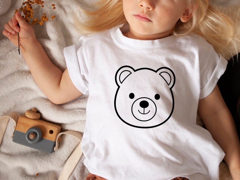 Może przedstawiać: Biały t-shirt z kr&oacute;tkim rękawem z czarnym obrysem głowy misia. Miś ma okrągłe czarne oczy i mały czarny nos. Koszulka jest noszona przez dziecko. Widoczny jest drewniany aparat fotograficzny.