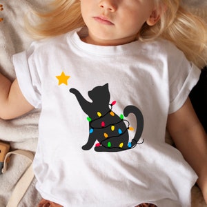 May include: White t-shirt with a black cat silhouette reaching for a yellow star. The cat is decorated with colourful Christmas lights. A festive design for the holiday season.