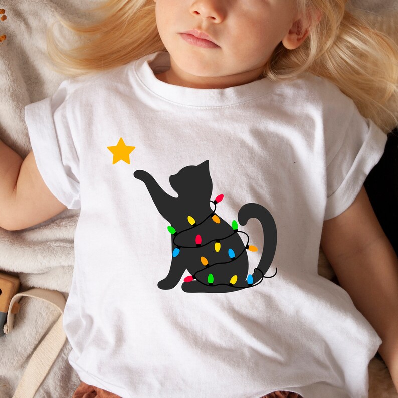 May include: White t-shirt with a black cat silhouette reaching for a yellow star. The cat is decorated with colourful Christmas lights. A festive design for the holiday season.