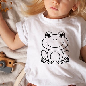 May include: White t-shirt featuring a black outline of a smiling frog. The frog has large eyes and a simple, friendly design. The shirt is worn by a child.