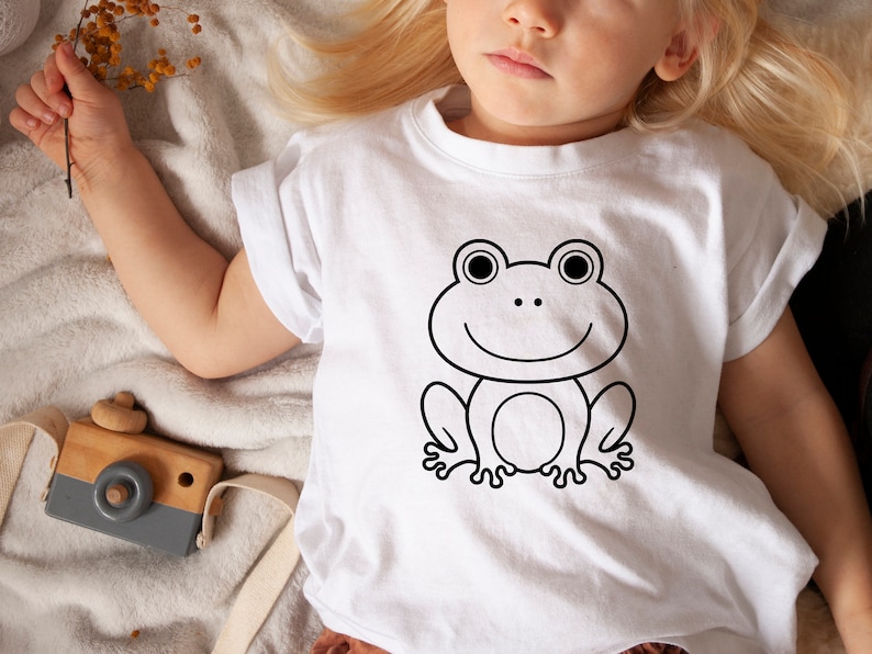 May include: White t-shirt featuring a black outline of a smiling frog. The frog has large eyes and a simple, friendly design. The shirt is worn by a child.