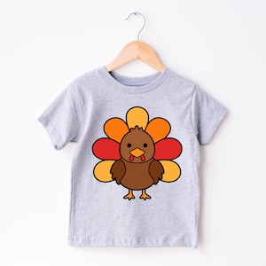May include: A grey short-sleeved t-shirt with a cartoon turkey design. The turkey is brown with a red wattle and a yellow and orange fan of feathers.