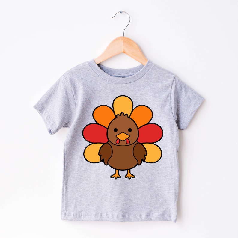 May include: A grey short-sleeved t-shirt with a cartoon turkey design. The turkey is brown with a red wattle and a yellow and orange fan of feathers.