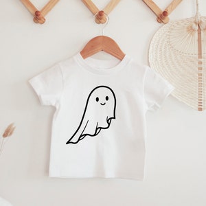 May include: A white t-shirt featuring a black outline of a smiling ghost. The ghost design has a flowing sheet shape. The t-shirt is displayed on a wooden hanger, suitable for Halloween or casual wear.