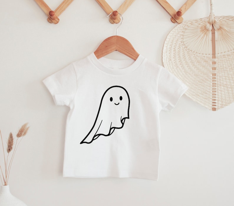 May include: A white t-shirt featuring a black outline of a smiling ghost. The ghost design has a flowing sheet shape. The t-shirt is displayed on a wooden hanger, suitable for Halloween or casual wear.