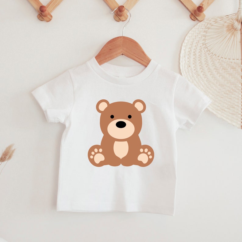 May include: White t-shirt featuring a brown teddy bear graphic. The bear has a black nose, brown ears, and tan paws. The shirt is hanging on a wooden hanger. The design is simple and suitable for children.