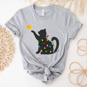 May include: A light grey t-shirt with a black cat silhouette reaching for a yellow star. The cat is wrapped in a string of colourful Christmas lights. The shirt is made of a soft material.