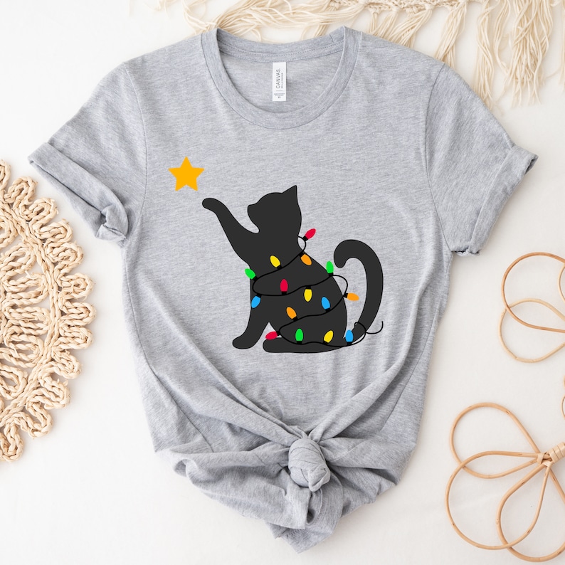 May include: A light grey t-shirt with a black cat silhouette reaching for a yellow star. The cat is wrapped in a string of colourful Christmas lights. The shirt is made of a soft material.