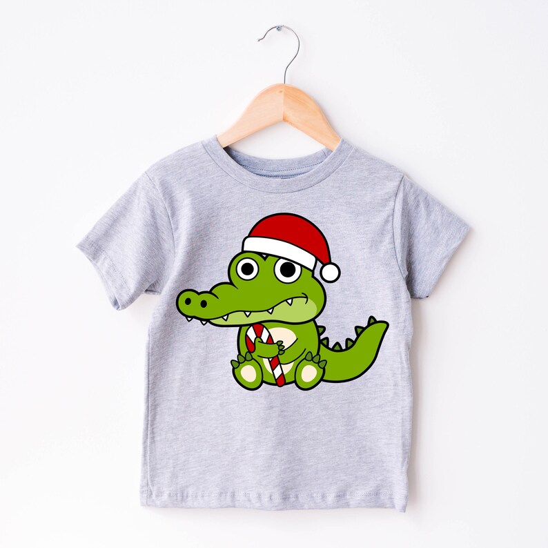 May include: A light gray t-shirt featuring a cartoon alligator wearing a Santa hat and holding a candy cane. The alligator is green with white eyes and a red and white striped candy cane. The shirt is hanging on a wooden hanger.