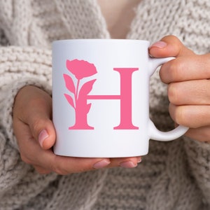 Pu&ograve; includere: Tazza in ceramica bianca con un motivo floreale rosa e la lettera "H". L'elemento floreale &egrave; a sinistra e la lettera "H" &egrave; a destra. La tazza &egrave; tenuta da una persona che indossa un maglione color crema.