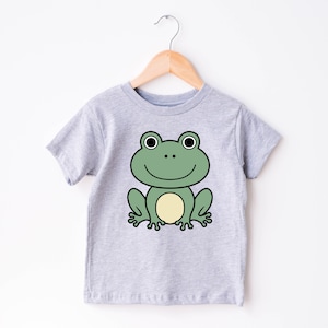 May include: A light gray t-shirt featuring a cartoon frog design. The frog is green with a yellow belly, black eyes, and a smiling mouth. The shirt is hanging on a wooden hanger.