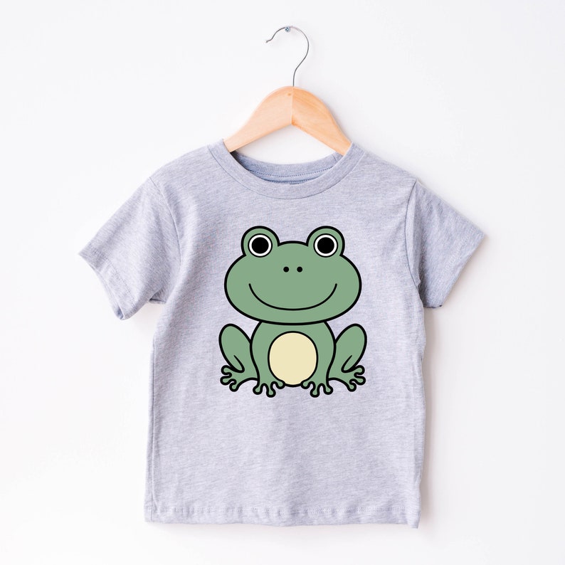 May include: A light gray t-shirt featuring a cartoon frog design. The frog is green with a yellow belly, black eyes, and a smiling mouth. The shirt is hanging on a wooden hanger.