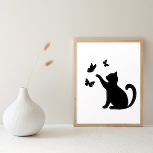 May include: A framed art print featuring a black silhouette of a cat reaching for butterflies. The cat is depicted in a playful pose, with its paw extended towards the fluttering insects. The artwork is set against a white background within a wooden frame.