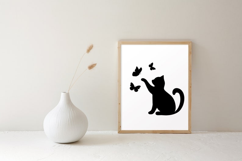 May include: A framed art print featuring a black silhouette of a cat reaching for butterflies. The cat is depicted in a playful pose, with its paw extended towards the fluttering insects. The artwork is set against a white background within a wooden frame.