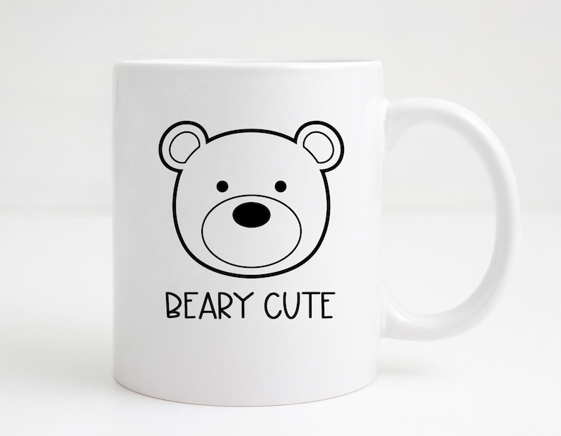 May include: White ceramic mug featuring a black outline illustration of a bear's face. The text "BEARY CUTE" is printed below the bear design. The mug has a curved handle.