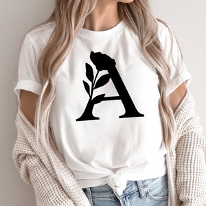 Pu&ograve; includere: T-shirt bianco con una grande lettera "A" nera con un design floreale. La grafica &egrave; centrata sulla parte anteriore della maglietta. La maglietta &egrave; realizzata in materiale morbido.