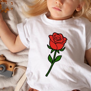 May include: White t-shirt featuring a large red rose graphic with green leaves and stem. The rose is centered on the shirt. The shirt is on a child.