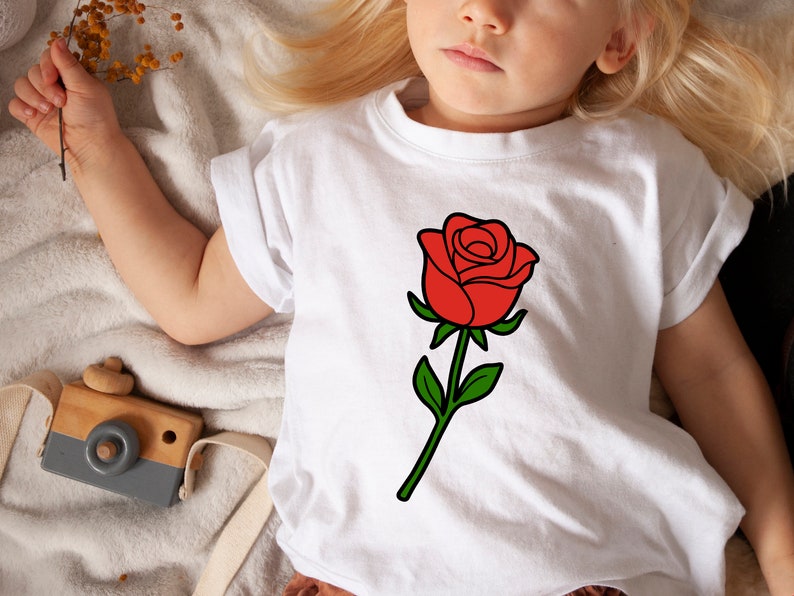 May include: White t-shirt featuring a large red rose graphic with green leaves and stem. The rose is centered on the shirt. The shirt is on a child.