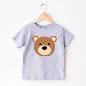 May include: A light gray t-shirt featuring a brown bear face design. The bear has a tan muzzle, brown ears, and black eyes. The shirt is hanging on a wooden hanger against a white background. This children's tee is perfect for everyday wear.