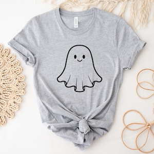 May include: A light grey t-shirt with a black outline of a smiling ghost. The ghost has a simple, smiling face. The shirt is made of a soft material and has rolled-up sleeves. The shirt is tied in a knot at the bottom.