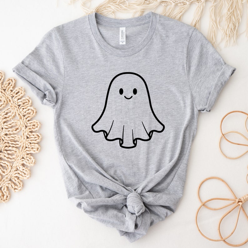 May include: A light grey t-shirt with a black outline of a smiling ghost. The ghost has a simple, smiling face. The shirt is made of a soft material and has rolled-up sleeves. The shirt is tied in a knot at the bottom.