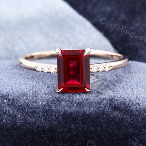 May include: A rose gold engagement ring with a red emerald cut gemstone and a band of small diamonds.