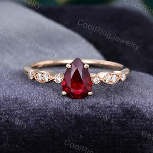 May include: A rose gold ring with a pear-shaped, deep red gemstone as the centerpiece. The band features small, clear accent stones set in a delicate pattern. The ring is displayed on a dark gray velvet surface.