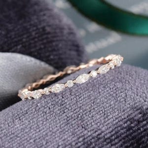May include: A delicate rose gold ring with a continuous band of small, sparkling diamonds. The ring features a mix of round and marquise-cut stones, set closely together. The ring is displayed on a dark gray velvet surface.