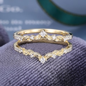 May include: A gold ring guard featuring a V-shaped chevron design with a textured leaf pattern and small round white stones set along the band. The ring has a polished finish and a symmetrical structure that creates a pointed centre peak.