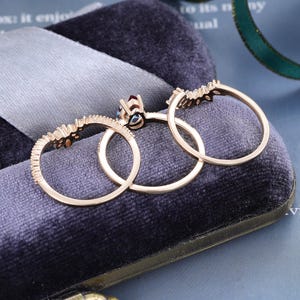 May include: A set of three rose gold rings featuring a central ring with a dark red gemstone and two matching curved bands adorned with small clear stones. The rings are arranged in a row, showcasing their thin metal bands and intricate settings.