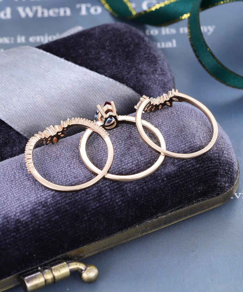 May include: A set of three rose gold rings featuring a central ring with a dark red gemstone and two matching curved bands adorned with small clear stones. The rings are arranged in a row, showcasing their thin metal bands and intricate settings.