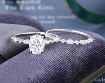 May include: A silver-toned engagement ring set featuring a central oval-cut stone flanked by marquise-cut stones on the band, accompanied by a matching wedding band with a series of marquise-cut stones set along the thin, polished metal band.
