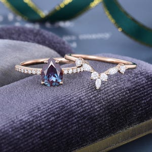 May include: A rose gold ring set featuring a pear-cut color-changing gemstone center stone flanked by small round diamonds on the band, paired with a matching V-shaped contour band adorned with marquise and round-cut white accent stones.