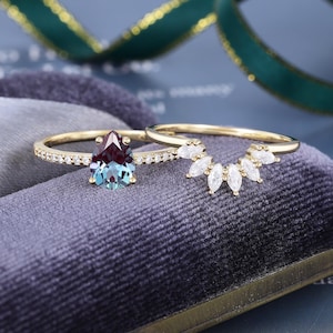 May include: A gold ring set featuring a pear-shaped color-changing alexandrite stone accented by small round diamonds on the band, paired with a curved gold band adorned with seven marquise-cut white stones arranged in a fan-like pattern.