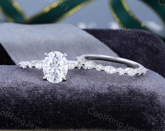 May include: A diamond engagement ring set featuring a large oval-cut center stone and a delicate band with smaller accent stones. The set includes a matching wedding band with marquise-shaped stones. The rings are displayed on a dark gray velvet surface.