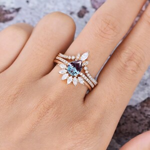 May include: A rose gold ring set featuring a pear-shaped color-changing center stone surrounded by marquise and round-cut white stones. The set includes a main ring and a curved band, both adorned with small pav&eacute;-set white stones along the bands.