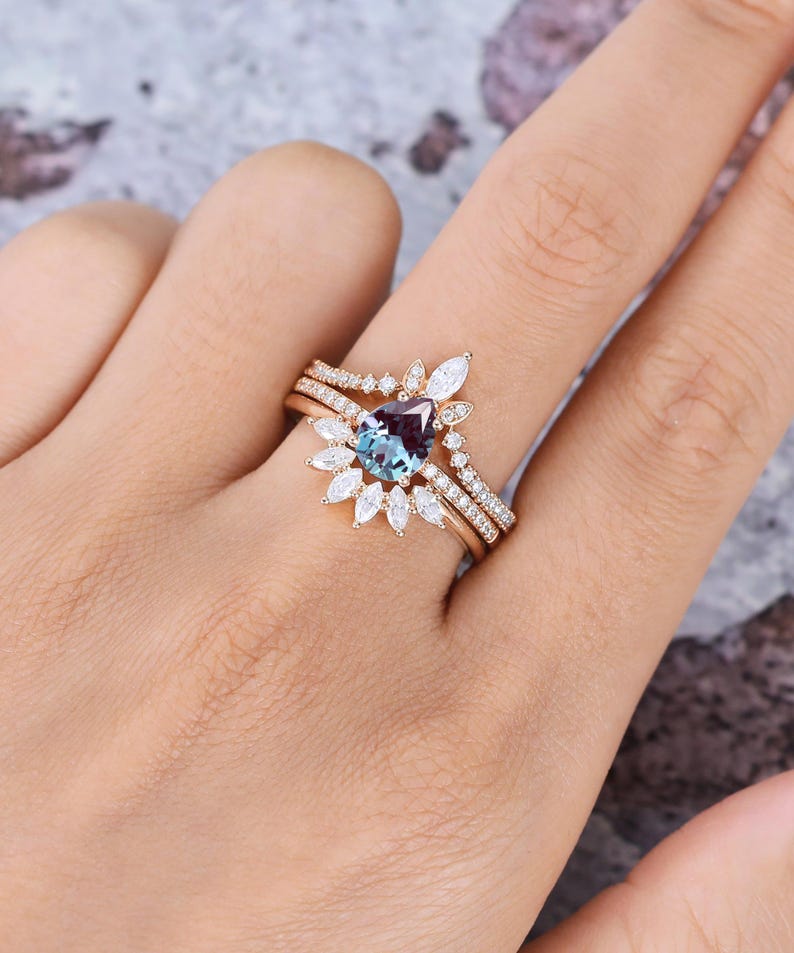 May include: A rose gold ring set featuring a pear-shaped color-changing center stone surrounded by marquise and round-cut white stones. The set includes a main ring and a curved band, both adorned with small pav&eacute;-set white stones along the bands.