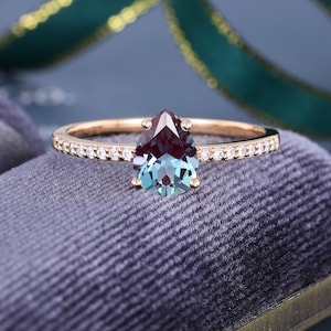 May include: A rose gold ring featuring a pear-shaped, color-changing alexandrite gemstone at the center, held by four prongs. The slender band is accented with a row of small, round, clear pavé-set diamonds extending along both sides.