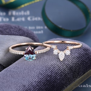 May include: A rose gold ring set featuring a pear-shaped color-changing gemstone flanked by small round diamonds on the band, paired with a matching curved band adorned with a central pear-shaped diamond and smaller round accent diamonds.