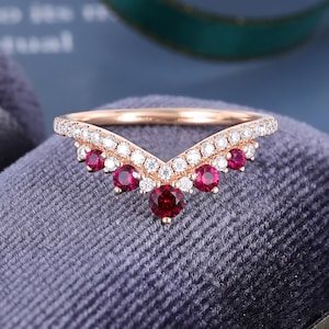 May include: A rose gold V-shaped ring featuring a row of small clear round stones along the band and five deep red round rubies set in a chevron pattern. The metal band has a polished finish and the stones are set in a prong setting.